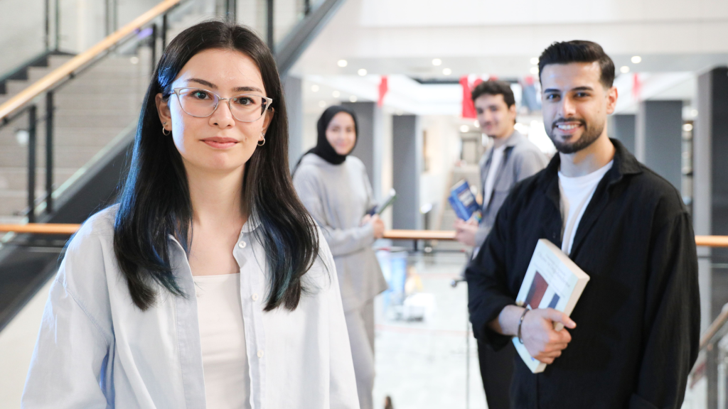 Female university student wearing glasses smiling at camera with diverse group of classmates holding books in modern campus building