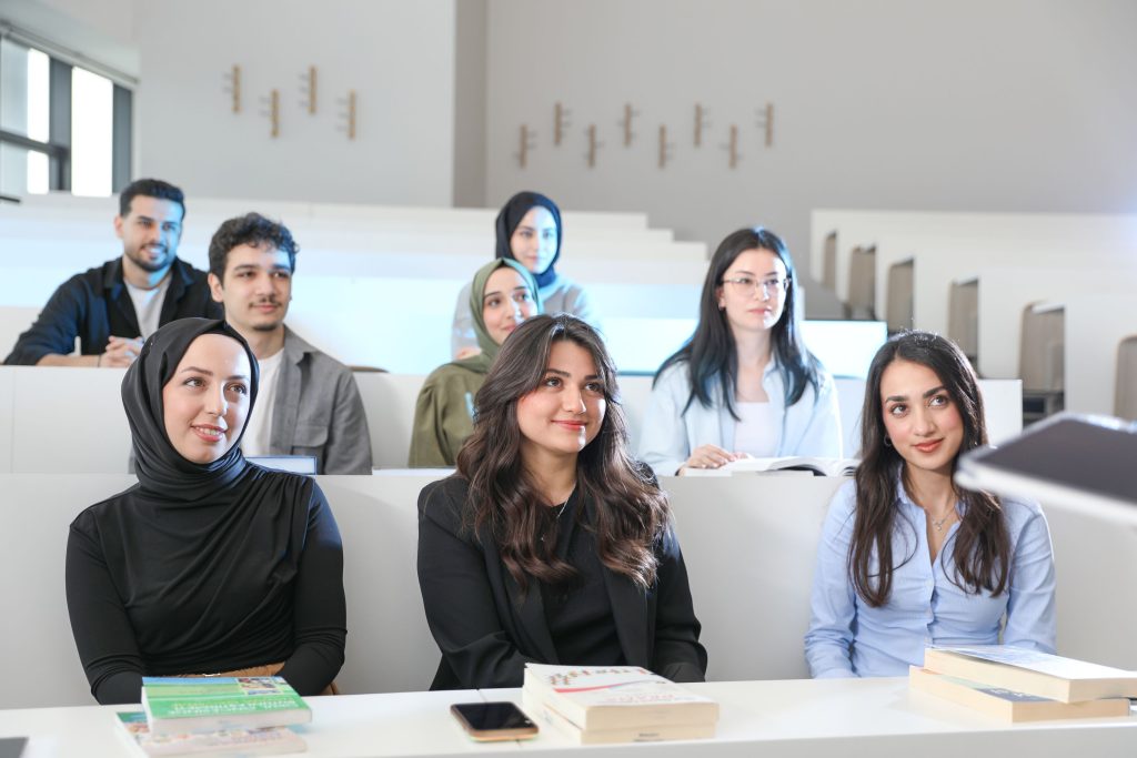 Group of students sitting in a lecture hall, smiling and listening attentively with books on the desks.
