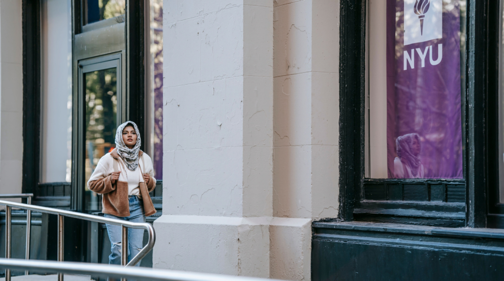 Young woman wearing a hijab walking outside a university building with an NYU banner visible in the window